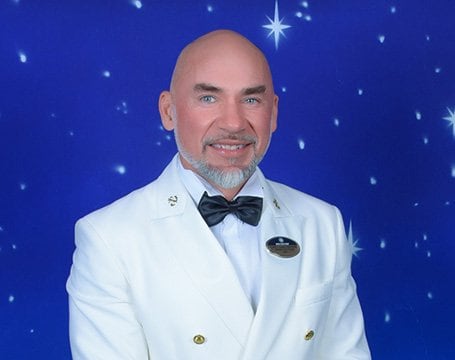 A crew member in a white uniform with a bow tie against a starry backdrop | MSC Cruises A crew member in a white uniform with a bow tie against a starry backdrop | MSC Cruises