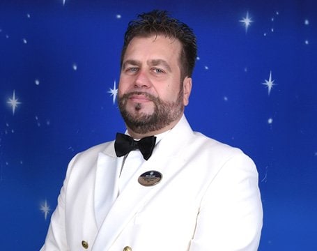Crew member in a white uniform with a bow tie against a starry blue background | MSC Cruises Crew member in a white uniform with a bow tie against a starry blue background | MSC Cruises