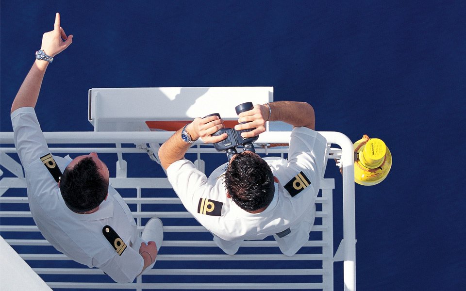 Two uniformed officers on a cruise ship deck, one using binoculars to scan the ocean | MSC Cruises Two uniformed officers on a cruise ship deck, one using binoculars to scan the ocean | MSC Cruises